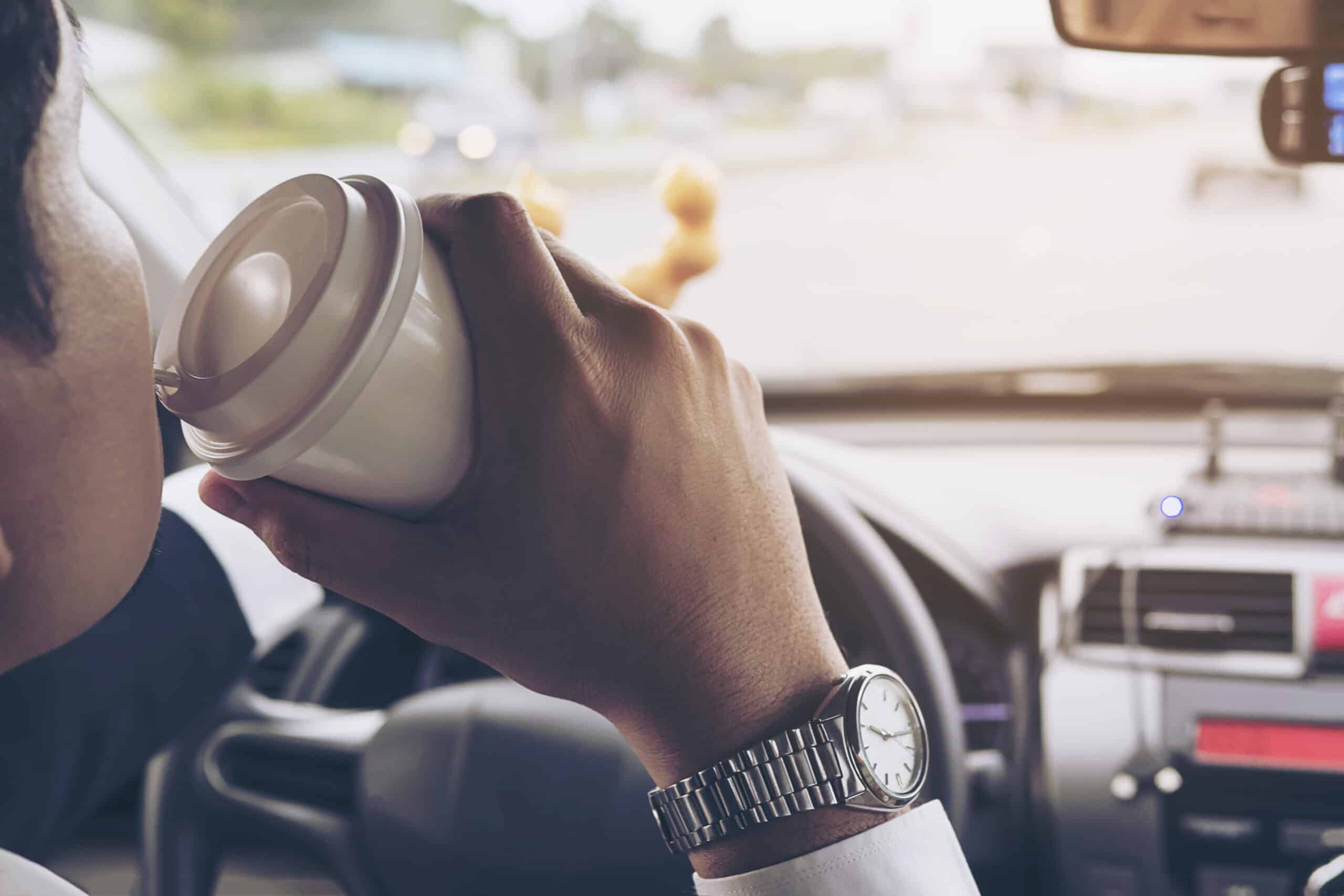 Man drinking coffee & holding a doughnut while driving car to show an example of distracted driving.