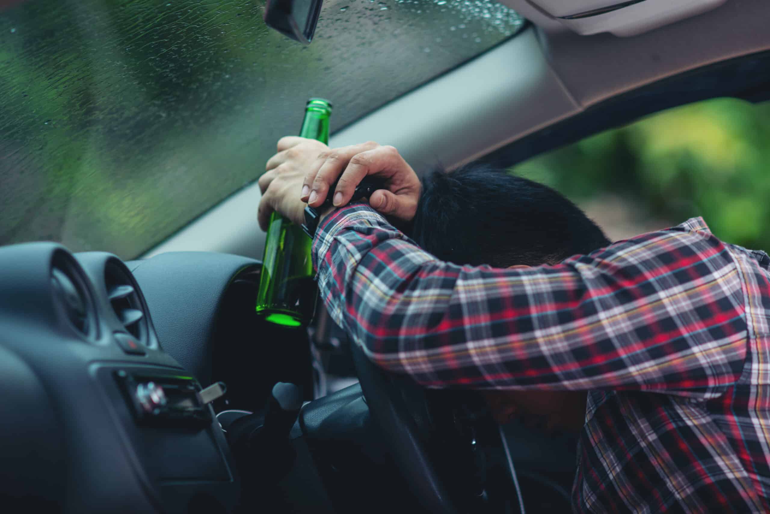 Picture shows a man with a beer bottle in hand and laying his head on the steering wheel, showing an example of how a drunk driving accident can occur